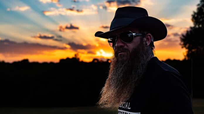 Cody Jinks, a man wearing sunglasses and a cowboy hat with a long beard, with a sunrise in the background