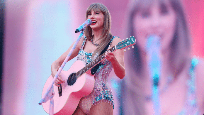 A woman with blonde hair in a pink and blue bodysuit, holding a pink guitar and smiling