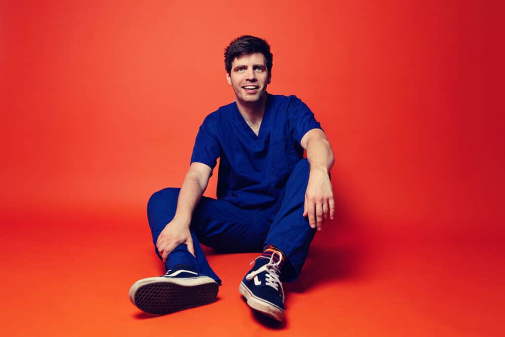 Comedian Ed Patrick is sat on the floor wearing his NHS uniform, set against a plain orange background