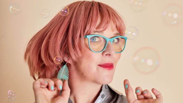Louise Leigh Distracted headshot. Louise has pink hair and turquoise glasses and matching nail varnish and jewellery. She is tilting her head to the side.